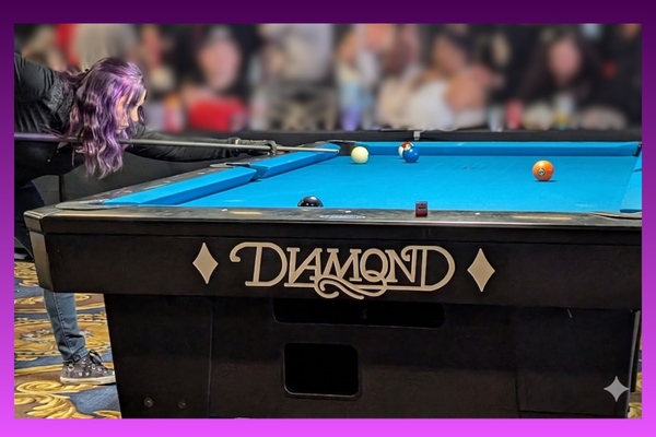 A side-view photograph of a woman with purple hair leaning over a professional black Diamond pool table, focused and preparing to take a shot. She is holding a pool cue, aligned with the white cue ball on the vibrant blue felt table. The surrounding room is brightly lit, showcasing a clean, high-end billiard setting that emphasizes focus and strategic planning.