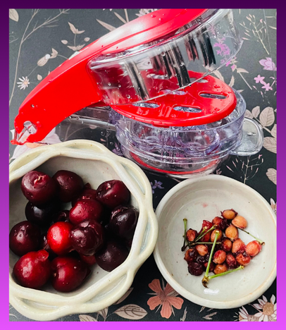 A top-down shot of a red and clear plastic multi-cherry pitter gadget resting on a dark floral tablecloth. To the left, a small light-colored ceramic bowl is filled with whole, dark red cherries. To the right, a matching smaller bowl contains a pile of discarded cherry pits and stems, showing the successful outcome of the tool. The entire image is framed with a deep purple border.
