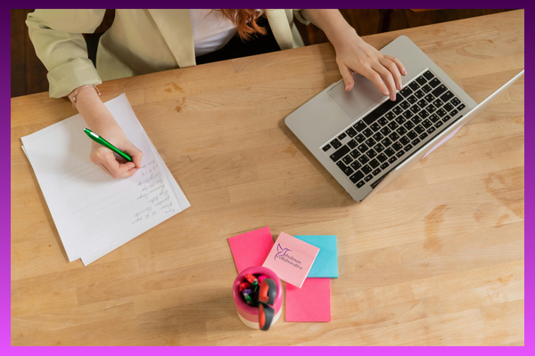 An overhead, flat lay view of a minimalist wooden office desk. A person's hands are visible: one typing on a silver laptop and the other writing a checklist with a green pen on a sheet of white paper. In the center are vibrant pink and blue sticky notes—including one featuring the Shulman Collaborative logo—next to a pink pen holder with scissors. The entire image is framed with a deep purple border.